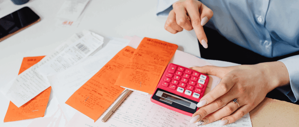 A imagem mostra uma pessoa fazendo contas em uma calculadora e analisando diversos papéis, representando a transformação que os registros empresariais terão no Brasil com o MAT, ou Módulo de Administração Tributária.