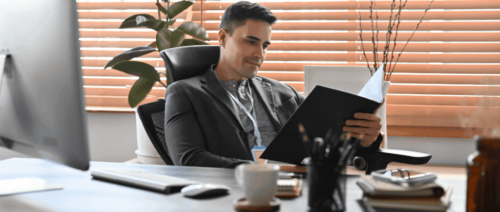 A imagem mostra um homem em seu escritório com uma mesa e, sobre ela, cadernos, lápis, uma caneca, um mouse, um teclado e um monitor. Ele está analisando os dados contábeis de seu negócio, representando um empresário que está verificando o que vai mudar para a sua empresa do Lucro Presumido com a Reforma Tributária.